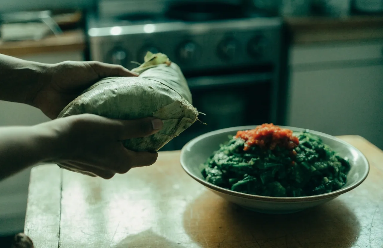 Hands lifting a leaf-wrapped parcel of sagu beside a bowl of cooked greens and sambal on a worn table